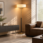 A modern floor lamp with a cylindrical white shade and slim metallic pole stands in a lit living room corner beside a brown leather armchair and a dark sideboard.